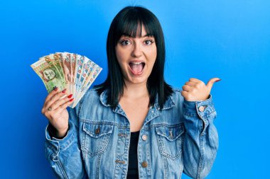 Young hispanic woman holding peruvian sol banknotes pointing thumb up to the side smiling happy with open mouth 