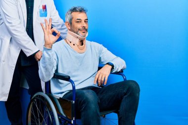 Handsome middle age man with grey hair on wheelchair wearing cervical collar smiling positive doing ok sign with hand and fingers. successful expression. 