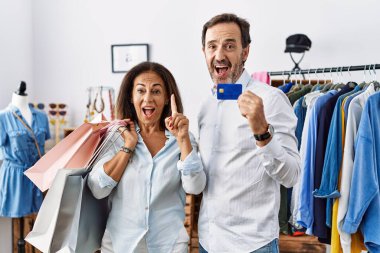 Hispanic middle age couple holding shopping bags and credit card pointing finger up with successful idea. exited and happy. number one. 