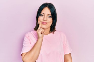 Young hispanic woman wearing casual pink t shirt looking confident at the camera with smile with crossed arms and hand raised on chin. thinking positive. 