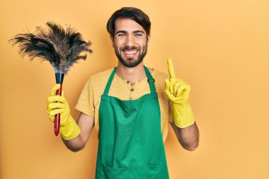 Young hispanic man wearing apron holding cleaning duster smiling with an idea or question pointing finger with happy face, number one 