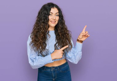 Gündelik kıyafetler giyen, gülümseyen ve kameraya bakan genç İspanyol kız iki eliyle ve parmaklarıyla kenarı işaret ediyor.. 