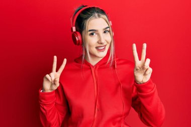 Young modern girl wearing gym clothes and using headphones smiling looking to the camera showing fingers doing victory sign. number two. 