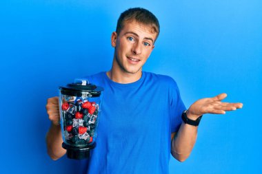 Young caucasian man making smoothie holding electric mixer celebrating achievement with happy smile and winner expression with raised hand 