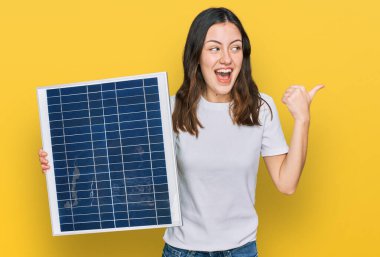 Genç ve güzel bir kız elinde fotovoltaik güneş paneli tutarak baş parmağını yukarı kaldırıyor ve ağzı açık gülümsüyor. 