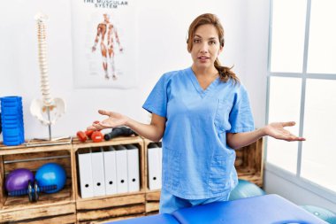 Middle age hispanic physiotherapist woman working at pain recovery clinic clueless and confused with open arms, no idea concept. 