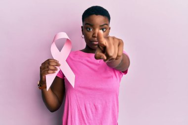 Young african american woman holding pink cancer ribbon pointing with finger to the camera and to you, confident gesture looking serious 