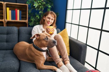 Kafkasyalı bir kadın kanepede köpeğiyle kahve içiyor.