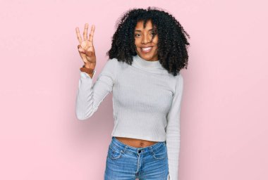 Genç Afro-Amerikan kızı, günlük kıyafetler giyip üç numaralı parmaklarıyla işaret ederken kendinden emin ve mutlu bir şekilde gülümsüyor.. 