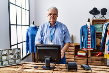 Orta yaşlı, gri saçlı, gülümseyen, kendine güvenen bir adam giyim mağazasında çalışıyor.