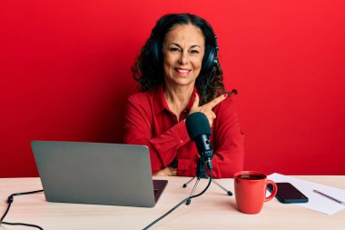Beautiful middle age woman working at radio studio cheerful with a smile of face pointing with hand and finger up to the side with happy and natural expression on face 