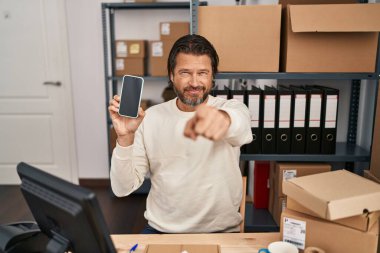 Handsome middle age man working at small business ecommerce holding smartphone pointing with finger to the camera and to you, confident gesture looking serious 