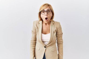 Middle age blonde business woman standing over isolated background in shock face, looking skeptical and sarcastic, surprised with open mouth 