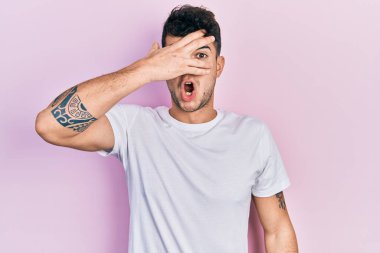 Günlük beyaz tişört giyen genç İspanyol adam şok içinde yüzünü ve gözlerini kapayarak parmaklarından korkuyla bakıyor. 