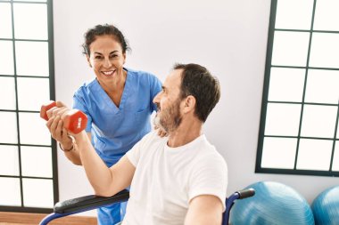 Orta yaşlı bir adam ve kadın, fizyoterapi kliniğinde tekerlekli sandalyede oturan dambılı kullanarak rehabilitasyon seansı yapıyorlar.