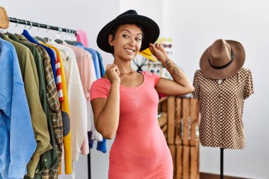 Young hispanic woman customer smiling confident choosing clothes wearing hat at clothing store