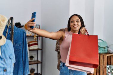 Genç Afrikalı Amerikalı bir kadın elbise mağazasında elinde çantalarla akıllı telefonuyla selfie çekiyor.