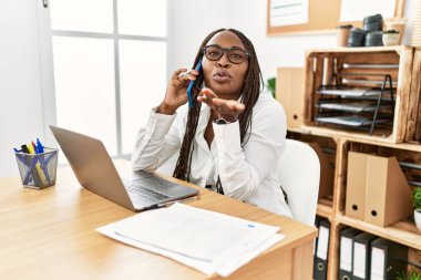 Black woman with braids working at the office speaking on the phone looking at the camera blowing a kiss with hand on air being lovely and sexy. love expression. 