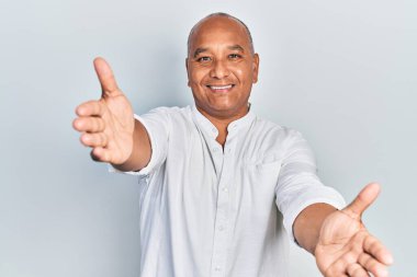 Orta yaşlı Latin bir adam, sıradan kıyafetler giyip kameraya bakıyor ve sarılmak için kollarını açmış gülümsüyor. Mutluluğu kucaklayan neşeli bir ifade.. 