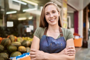 Meyve dükkanında önlük giyerek mutlu gülümseyen genç sarışın kadın.