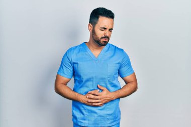 Handsome hispanic man with beard wearing blue male nurse uniform with hand on stomach because indigestion, painful illness feeling unwell. ache concept. 