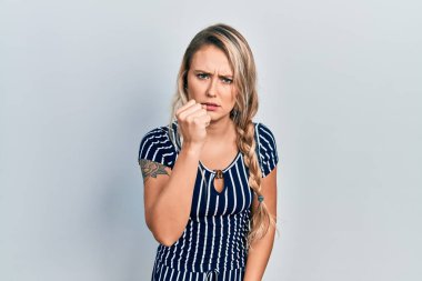 Beautiful young blonde woman wearing casual striped dress angry and mad raising fist frustrated and furious while shouting with anger. rage and aggressive concept. 