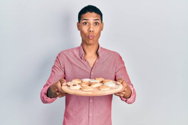Young african american guy holding tray with cake sweets making fish face with mouth and squinting eyes, crazy and comical. 