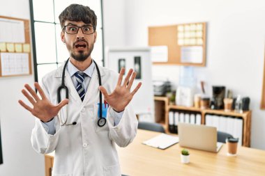 Hispanic man with beard wearing doctor uniform and stethoscope at the office afraid and terrified with fear expression stop gesture with hands, shouting in shock. panic concept. 