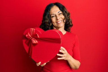 Middle age hispanic woman holding valentine day gift looking positive and happy standing and smiling with a confident smile showing teeth 