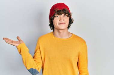 Handsome young man wearing wool hat smiling cheerful presenting and pointing with palm of hand looking at the camera. 