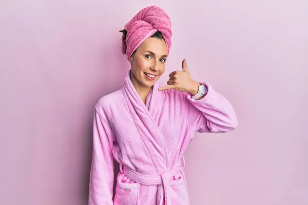 Young blonde woman wearing shower towel cap and bathrobe smiling doing phone gesture with hand and fingers like talking on the telephone. communicating concepts.