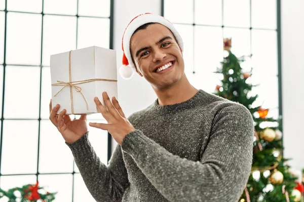 Young Hispanic Man Holding Gift Standing Christmas Tree Home — Stok fotoğraf