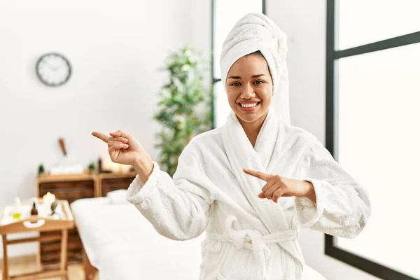 Young brunette woman wearing towel and bathrobe standing at beauty center smiling and looking at the camera pointing with two hands and fingers to the side.