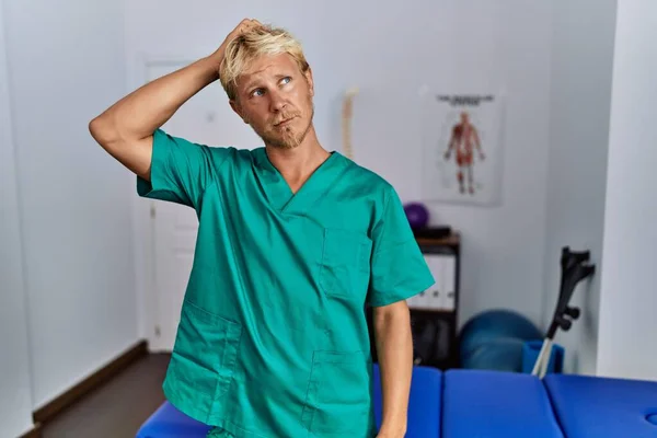 Young Blond Man Wearing Physiotherapist Uniform Standing Clinic Confuse Wonder — Stok fotoğraf