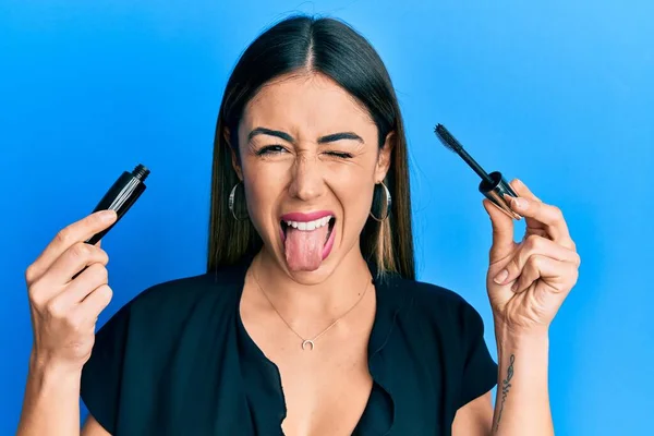 Young Hispanic Woman Holding Eyelash Mascara Sticking Tongue Out Happy — Stockfoto