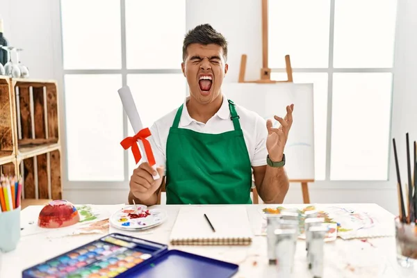 Young Hispanic Man Art Studio Holding Degree Crazy Mad Shouting — стоковое фото