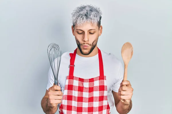 Young hispanic man with modern dyed hair wearing cook apron holding baker whisk and spoon making fish face with mouth and squinting eyes, crazy and comical.