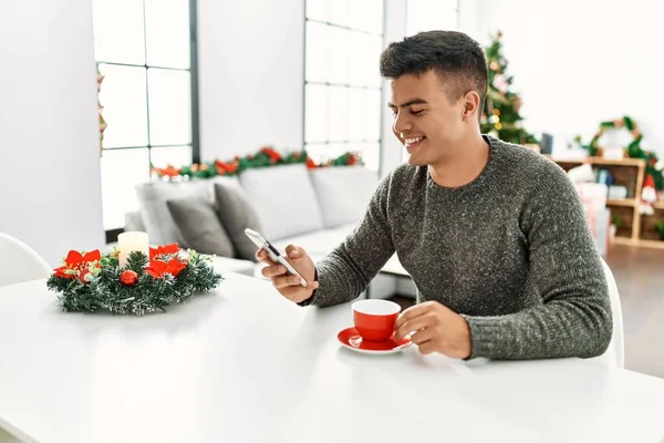 Young Hispanic Man Drinking Coffee Using Smartphone Sitting Christmas Tree — 图库照片