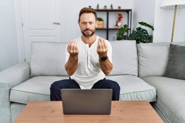 Middle age man using laptop at home doing money gesture with hands, asking for salary payment, millionaire business 