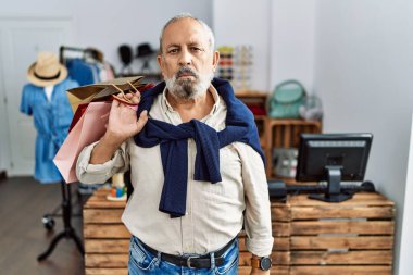 Handsome senior man holding shopping bags at boutique shop relaxed with serious expression on face. simple and natural looking at the camera. 