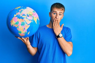 Young caucasian man holding world ball covering mouth with hand, shocked and afraid for mistake. surprised expression 
