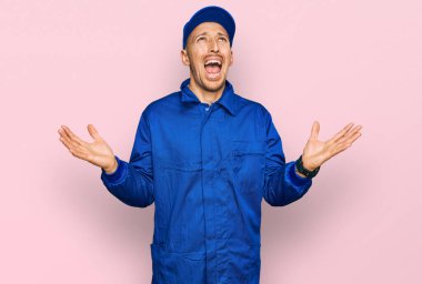 Bald man with beard wearing builder jumpsuit uniform crazy and mad shouting and yelling with aggressive expression and arms raised. frustration concept. 