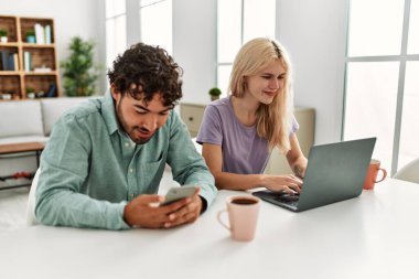 Laptop ve akıllı telefon kullanan genç bir çift evde kahve içiyor..