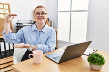 Ofiste çalışan genç kızıl saçlı kadın bilgisayarlı dizüstü bilgisayar kullanıyor. Ellerinde büyük boyut işareti, ölçü sembolü var. Kameraya bakıp gülümsüyor. ölçüm kavramı. 