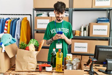 Young hispanic man wearing volunteer uniform usong smartphone at charity center.