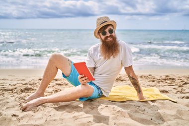 Genç kızıl saçlı adam plajda havlunun üzerinde oturuyor..