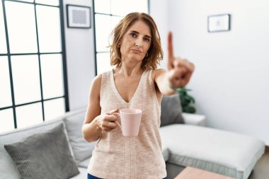 Orta yaşlı bir kadın evde kahve içiyor, parmağını kaldırmış ve kızgın bir ifadeyle işaret ediyor, hiçbir hareket göstermiyor. 