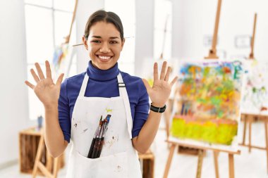 Young brunette woman at art studio showing and pointing up with fingers number ten while smiling confident and happy. 