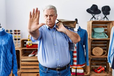 Gri saçlı yaşlı adam perakende mağazasında alışveriş torbaları tutuyor, elinde açık elle dur işareti yapıyor, ciddi ve kendinden emin bir ifadeyle, savunma jesti yapıyor. 