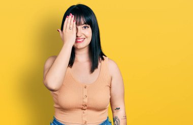 Latin kökenli genç ve iri yarı bir kadın günlük kıyafetler giyiyor. Bir gözünü eliyle kapatıyor. Yüzünde kendine güvenen bir gülümseme ve sürpriz bir duygu var.. 